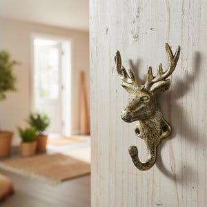 Könnte beinhalten: Ein bronzefarbener Wandhaken in Form eines Hirschkopfes. Der Haken ist auf einer hellen Holzoberfläche befestigt. Der Hirschkopf hat detaillierte Geweihe und einen gebogenen Haken zum Aufhängen von Gegenständen.