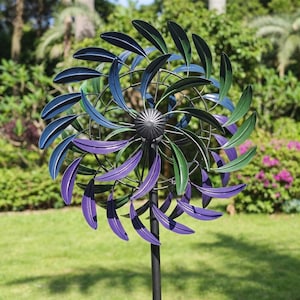 Könnte beinhalten: Ein dekorativer Metall-Windspinner für den Garten. Die Flügel sind in Blau-, Grün- und Lilatönen lackiert. Der Windspinner ist auf einer schwarzen Metallstange montiert und vor dem Hintergrund von grünem Laub und Gras platziert.