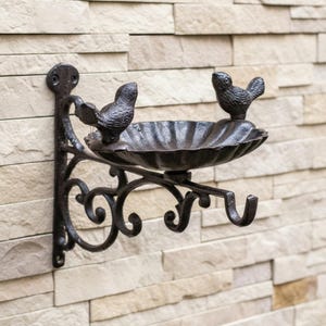 Puede incluir: Un bebedero decorativo de metal marrón oscuro con dos figuras de pájaros posadas en el borde. El bebedero está montado en una pared con un soporte decorativo y un fondo de piedra texturizada. El bebedero tiene un borde festoneado.