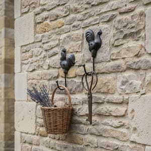 May include: A stone wall with decorative metal hooks. Two rooster-shaped hooks and a key-shaped hook are visible. A woven basket filled with lavender and yarn hangs from one hook. The scene evokes a rustic, country style.