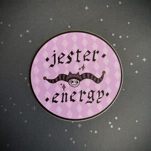 May include: A circular sticker with a light purple diamond pattern background. The text jester energy is printed in black gothic font. Between the words is a graphic of a jester hat with a face, featuring small gold star accents.