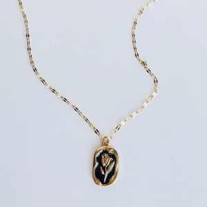Op de afbeelding: Een goudkleurige ketting met een rechthoekige hanger met een zwarte achtergrond en een wit bloemmotief. De ketting heeft een delicaat, plat schakelontwerp. De hanger is goudkleurig omlijnd.
