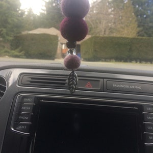 Op de afbeelding: Een auto-interieur met een achteruitkijkspiegelhanger met drie vilten ballen in tinten paars en bordeauxrood, een zilveren hanger en een bladvormige hanger. De hanger hangt aan de achteruitkijkspiegel.