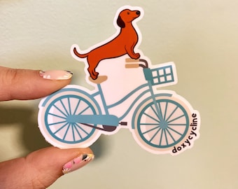 Peut inclure: Un autocollant représentant un teckel de dessin animé sur un vélo bleu clair avec un panier. Le teckel est marron et le vélo a des pneus blancs et un cadre turquoise. Le texte "doxycline" est imprimé sur l'autocollant.