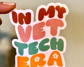 Op de afbeelding: Een sticker met de tekst "In My Vet Tech Era" in kleurrijke, afgeronde letters. De letters zijn in tinten oranje, roze, groen en bruin. Er zitten kleine witte bloemetjes op de "E" en "A" van "Era".