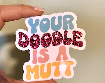 Op de afbeelding: Een sticker met een roze luipaardprint achtergrond en de tekst "YOUR DOODLE IS A MUTT" in kleurrijke, retro-stijl letters.