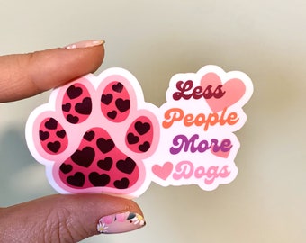 Può includere: Adesivo rosa a forma di impronta di zampa con un motivo di cuori rosa. L'adesivo dice "Meno persone Più cani" con un cuore rosa prima di "Cani".