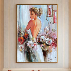 May include: An oil painting depicts a woman in a white dress, her back to the viewer, surrounded by flowers. The artwork is framed in gold and hangs above a bed with decorative pillows. The color palette includes white, red, pink, and gold.