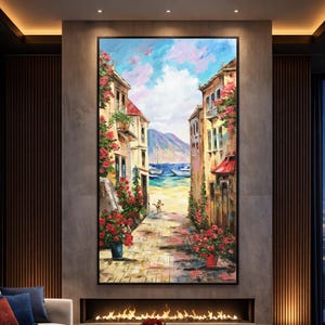 May include: Large framed oil painting depicting a coastal street scene. Buildings with red awnings and blooming red flowers line a cobblestone path leading to the sea. The sky is a mix of blue and pink hues, with boats visible in the distance.