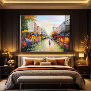 May include: Oil painting of a European street scene with cafes, shops, and pedestrians. The artwork features a vibrant color palette with warm tones, a golden frame, and is displayed above a bed with decorative pillows and a bench.