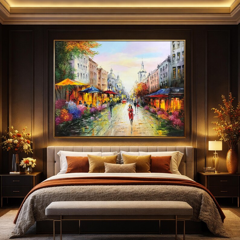 May include: Oil painting of a European street scene with cafes, shops, and pedestrians. The artwork features a vibrant color palette with warm tones, a golden frame, and is displayed above a bed with decorative pillows and a bench.