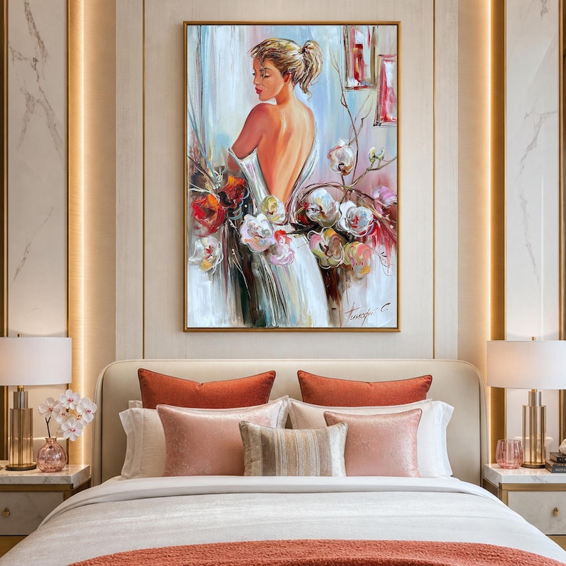 May include: An oil painting depicts a woman in a white dress, her back to the viewer, surrounded by flowers. The artwork is framed in gold and hangs above a bed with decorative pillows. The color palette includes white, red, pink, and gold.