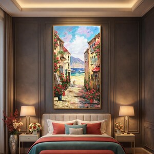 May include: A framed oil painting depicts a European coastal street scene. Buildings with red awnings and flower boxes line a cobblestone path leading to the sea. The sky is blue with fluffy white clouds. The painting is displayed above a bed with red and blue pillows.