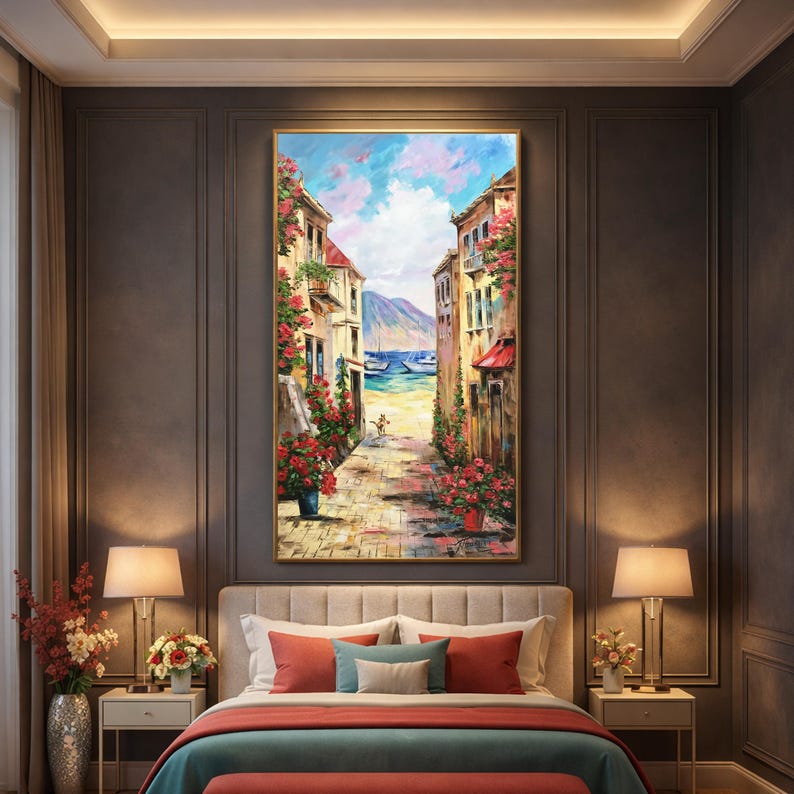 May include: A framed oil painting depicts a European coastal street scene. Buildings with red awnings and flower boxes line a cobblestone path leading to the sea. The sky is blue with fluffy white clouds. The painting is displayed above a bed with red and blue pillows.