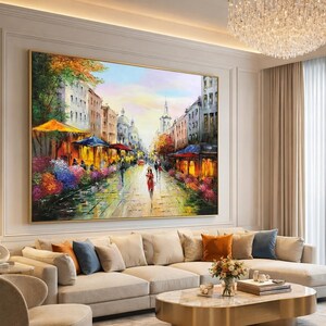 May include: Large framed oil painting depicting a European street scene. The artwork features a woman in a red dress walking down a cobblestone street lined with buildings, cafes, and colorful flowers. The painting has a gold frame.
