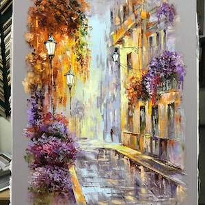 May include: An impressionistic oil painting of a European street scene. The artwork depicts a cobblestone street with buildings on either side, adorned with flowers and street lamps. The color palette includes shades of yellow, orange, purple, and gray.