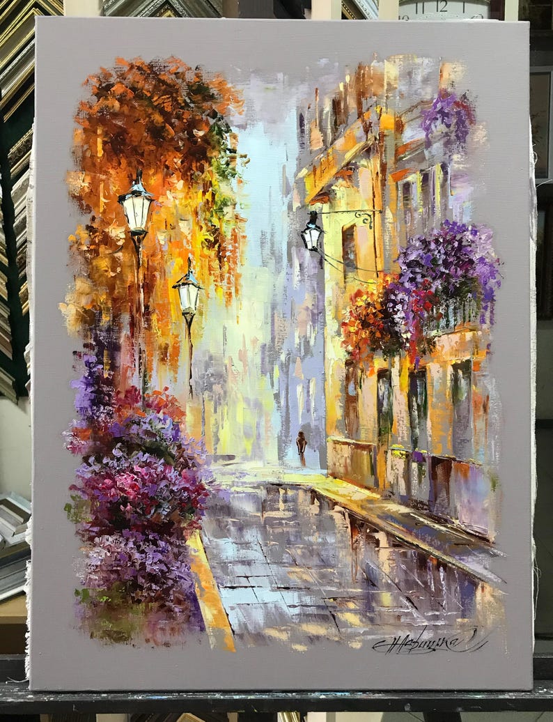 May include: An impressionistic oil painting of a European street scene. The artwork depicts a cobblestone street with buildings on either side, adorned with flowers and street lamps. The color palette includes shades of yellow, orange, purple, and gray.