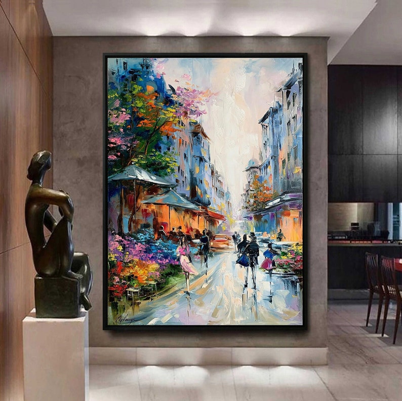 May include: A colorful oil painting depicting a street scene in a European city. The painting features buildings with colorful facades, a cobblestone street, and people walking. There are trees and flowers in the foreground, and the sky is a bright blue.