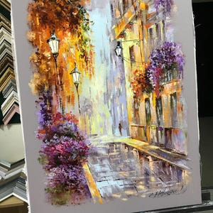 May include: An impressionistic oil painting of a European street scene. The artwork depicts a cobblestone street with buildings, street lamps, and colorful flowers. The color palette includes shades of orange, yellow, purple, and gray, creating a vibrant and atmospheric piece.