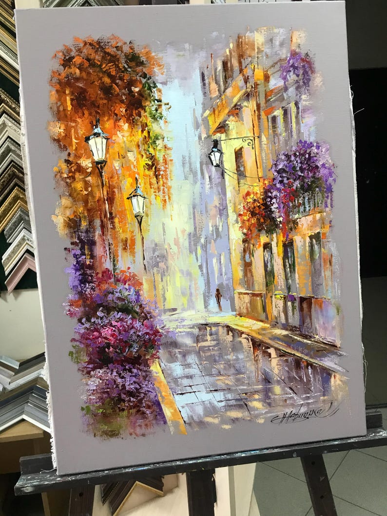 May include: An impressionistic oil painting of a European street scene. The artwork depicts a cobblestone street with buildings, street lamps, and colorful flowers. The color palette includes shades of orange, yellow, purple, and gray, creating a vibrant and atmospheric piece.