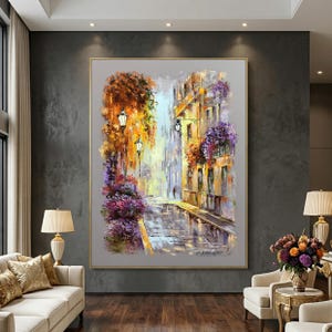 May include: An oil painting depicts a European street scene with buildings, street lamps, and colorful flowers. The artwork features a muted color palette with shades of yellow, purple, and gray. The painting is framed in a gold-colored frame and hangs on a dark gray wall.