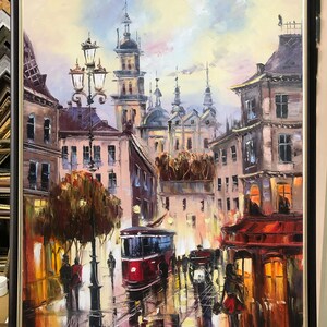 May include: Oil painting depicting a European city street scene with a red tram, buildings, and streetlights. The artwork is framed in a black frame. The painting has a warm colour palette with reflections on the wet street.