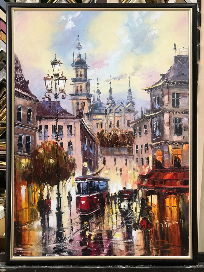 May include: Oil painting depicting a European city street scene with a red tram, buildings, and streetlights. The artwork is framed in a black frame. The painting has a warm colour palette with reflections on the wet street.