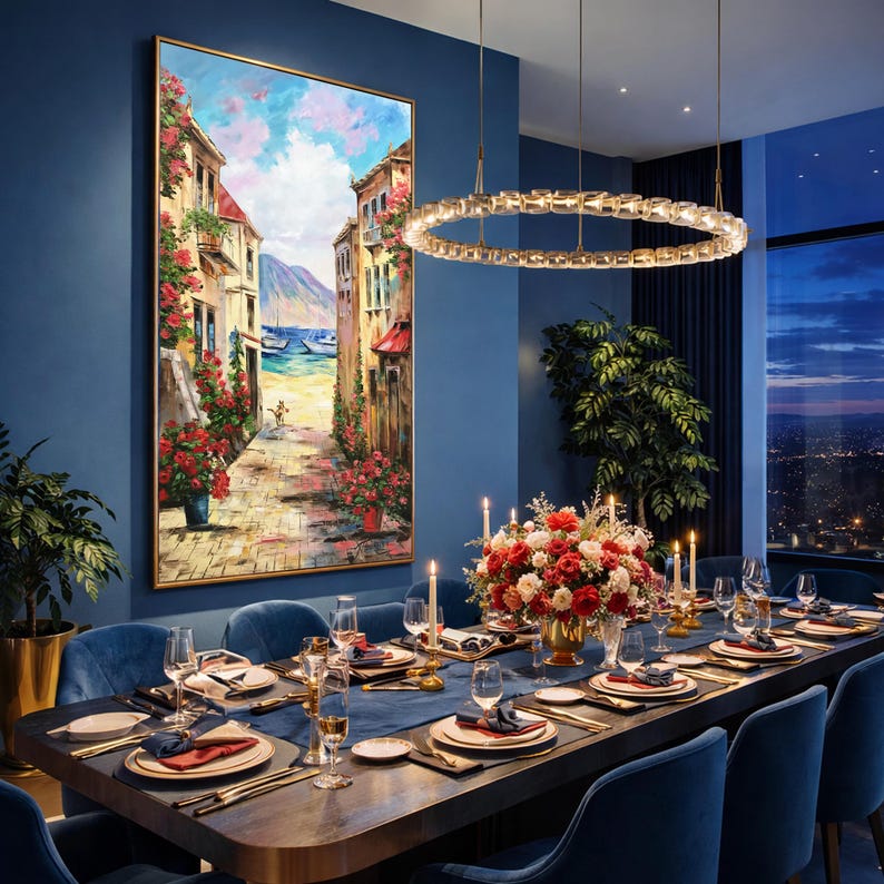 May include: A dining room with a large painting of a European street scene. The table is set for a formal dinner with blue velvet chairs, a floral centerpiece, and a modern chandelier. The painting features a cobblestone street, buildings, and boats.