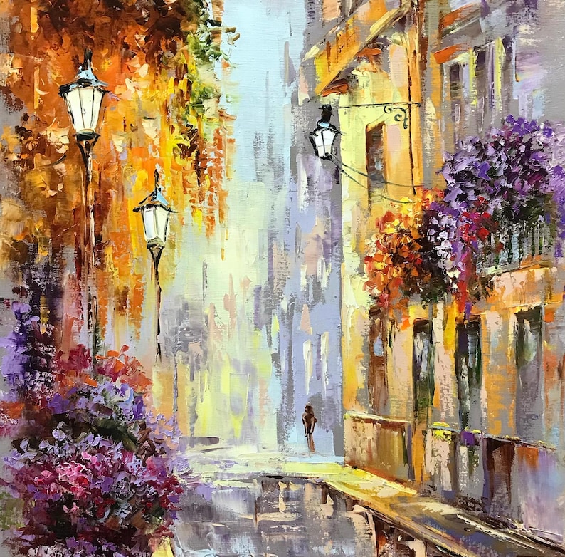 May include: An impressionistic oil painting of a European street scene. The artwork features a narrow street with buildings on either side, adorned with hanging flowers and street lamps. The color palette includes warm yellows, oranges, and purples.