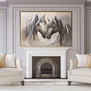 May include: An oil painting of two white horses with flowing manes, set in a gold frame. The artwork hangs above a white fireplace, flanked by two cream-colored armchairs with gold pillows. The room has gray walls and gold sconces.