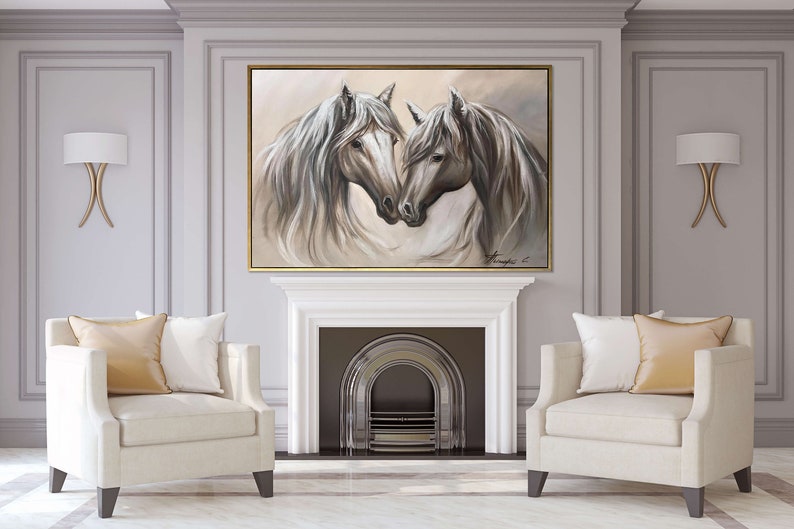 May include: An oil painting of two white horses with flowing manes, set in a gold frame. The artwork hangs above a white fireplace, flanked by two cream-colored armchairs with gold pillows. The room has gray walls and gold sconces.