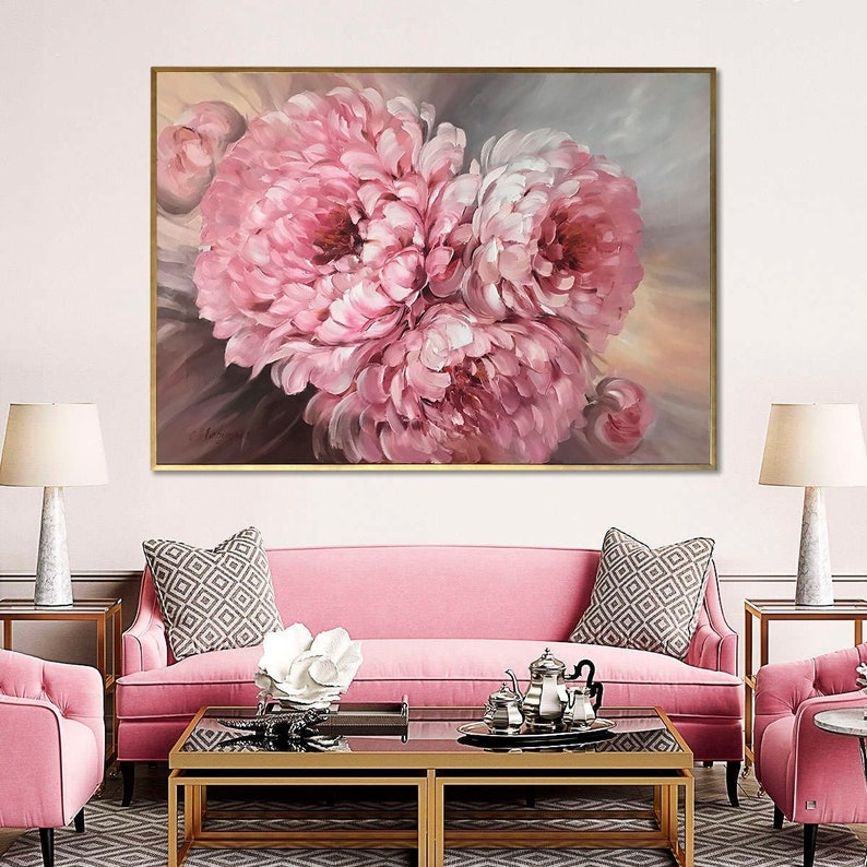 May include: A painting of pink peonies in a gold frame. The painting is hanging on a wall above a pink sofa with gold legs. The sofa has two throw pillows with a geometric pattern. There is a coffee table in front of the sofa with a glass top and gold legs. There are two gold end tables on either side of the sofa.