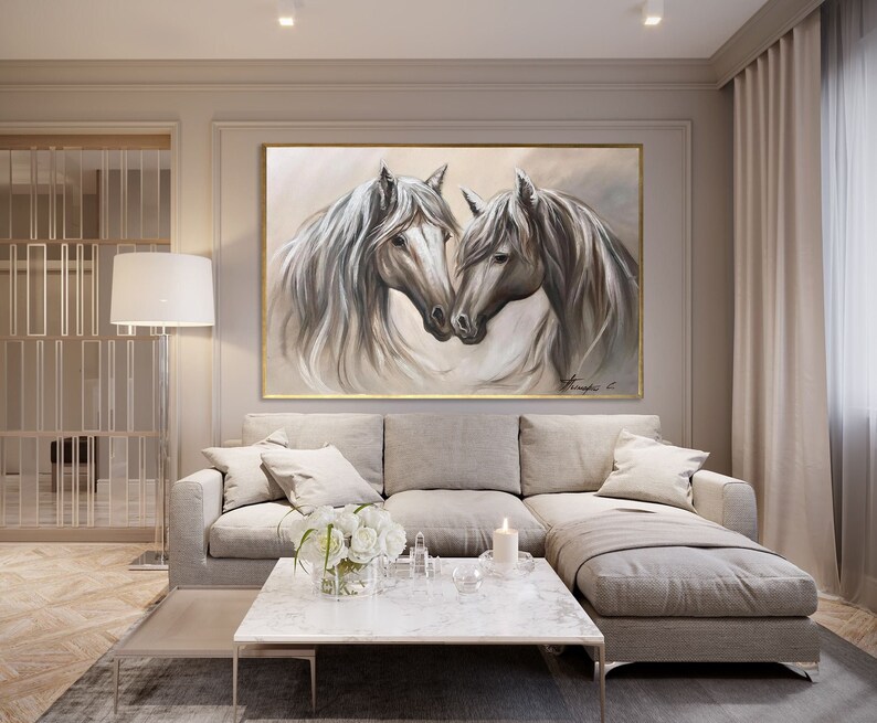 May include: A large framed painting depicts two white horses with flowing manes, heads touching. The artwork is in shades of gray and white, with a gold frame. The painting hangs above a gray sectional sofa in a modern living room setting.