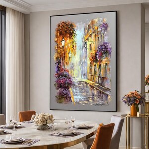 May include: An oil painting depicts a European street scene with buildings, streetlights, and colorful flowers. The artwork features a palette of warm colors, including orange, yellow, and purple, with a reflective street surface. The painting is framed in a black border.