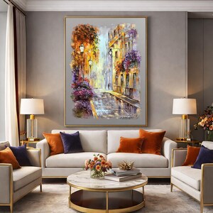 May include: An oil painting of a European street scene with buildings, streetlights, and colorful flowers. The artwork is framed in gold and hangs above a white sofa with orange and navy blue throw pillows. Two matching lamps flank the painting.
