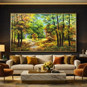 May include: A large framed landscape painting depicting a forest scene with a path. The artwork features trees with green, yellow, and orange foliage, suggesting an autumn setting. The painting hangs above a beige sofa with colorful throw pillows.