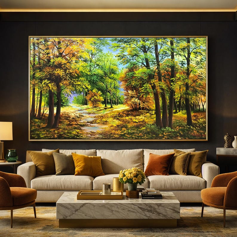 May include: A large framed landscape painting depicting a forest scene with a path. The artwork features trees with green, yellow, and orange foliage, suggesting an autumn setting. The painting hangs above a beige sofa with colorful throw pillows.