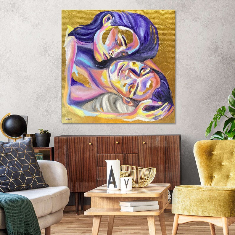 May include: A colorful painting depicts two figures embracing. The figures have closed eyes and are painted in shades of purple, yellow, pink, and blue. The background is gold. The artwork is displayed in a living room setting.