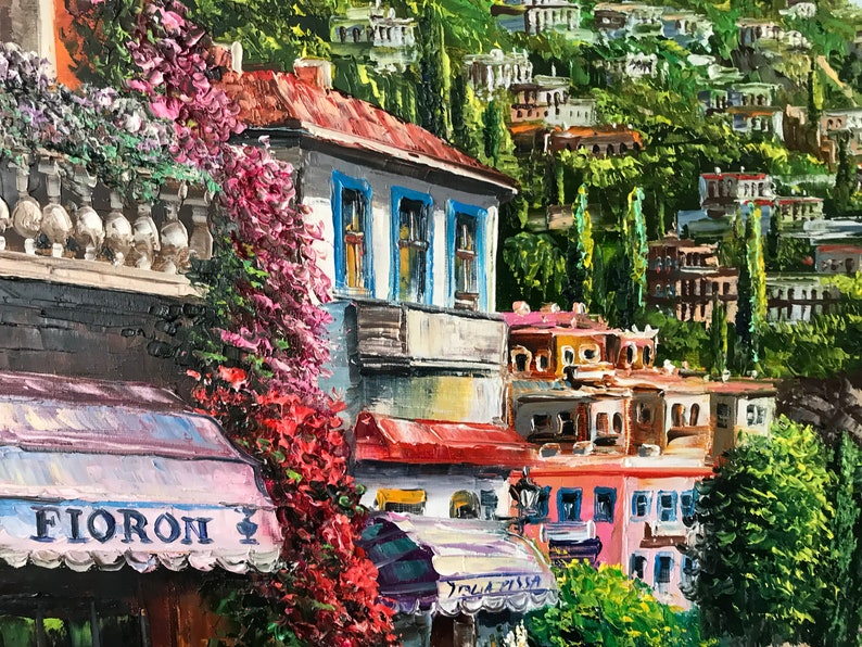 May include: A painting of a colorful, European-style street scene with a cafe awning that reads "FIORON". The cafe is surrounded by lush greenery and colorful buildings.