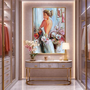 May include: An elegant dressing room featuring a framed painting of a woman with flowers, a marble-topped console table with gold legs, and a gold lamp. The room has built-in closets with hanging clothes and a neutral color palette.