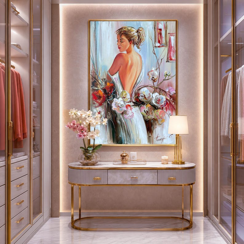 May include: An elegant dressing room featuring a framed painting of a woman with flowers, a marble-topped console table with gold legs, and a gold lamp. The room has built-in closets with hanging clothes and a neutral color palette.