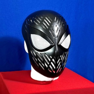 Peut inclure: Un masque en plastique noir avec un design détaillé et une protection oculaire en maille. Le masque est sur une surface rouge.