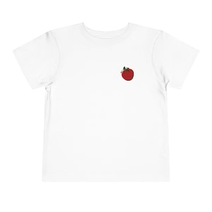 May include: White toddler t-shirt featuring a small, embroidered red tomato with green leaves on the left chest. The short-sleeved shirt is made of a soft, lightweight fabric, perfect for casual wear.