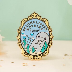 Könnte beinhalten: Goldfarbene Emaille-Anstecknadel mit einem weißen Seekuh in einer blauen und grünen Ozeanszene. Die Anstecknadel hat einen goldenen Rahmen mit dem Text "Accomplished Nothing Today" außen herum.