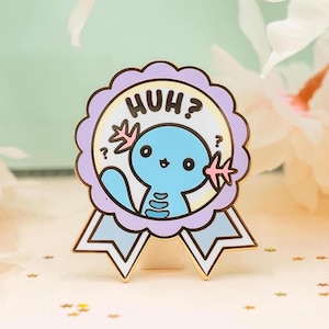 May include: A circular enamel pin shaped like a ribbon award with a light purple scalloped border. The centre features a light blue axolotl character with the text "HUH?" above it, accompanied by small question marks and pink branch-like accents.