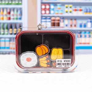 May include: A clear acrylic keychain shaped like a bento box, featuring a red border and internal compartments. Inside are illustrations of rice with a plum, fried shrimp, and egg. A white price tag displays the text 弁当 ¥900 and a barcode.