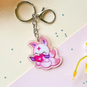 May include: A clear acrylic keychain featuring a pink and white cartoon fox holding a heart. The keychain has a silver-colored clasp and chain. The background includes pink and yellow paper, flowers, and a small notepad.