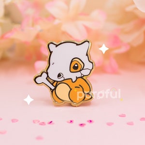 May include: A gold-colored enamel pin featuring a cartoon Cubone character. The pin has a white skull head, a tan body, and a bone accessory. The pin is set against a soft pink background with floral accents and heart-shaped confetti.