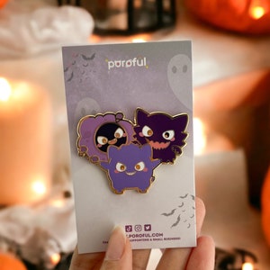 May include: A set of three enamel pins featuring purple cartoon ghost characters with gold trim. The pins are on a gray card with the brand name "poroful" and social media icons. The background includes pumpkins and candles.