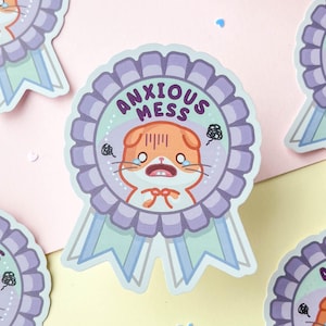 May include: Five stickers with a cartoon illustration of a sad orange cat with the text "ANXIOUS MESS" on a purple and light blue ribbon. The stickers are on a light pink and yellow background.